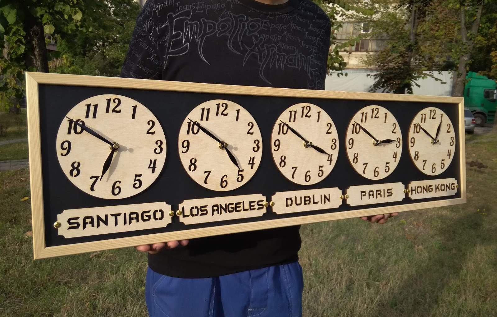 A person holding a personalized wooden wall clock with multiple time zones displayed in a long horizontal format, with city names below each clock face.