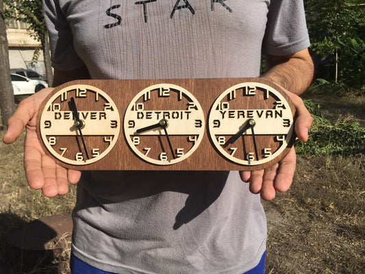 A person holding a wooden wall clock with three dial faces displaying 'DENVER', 'DETROIT', and 'YEREVAN'.